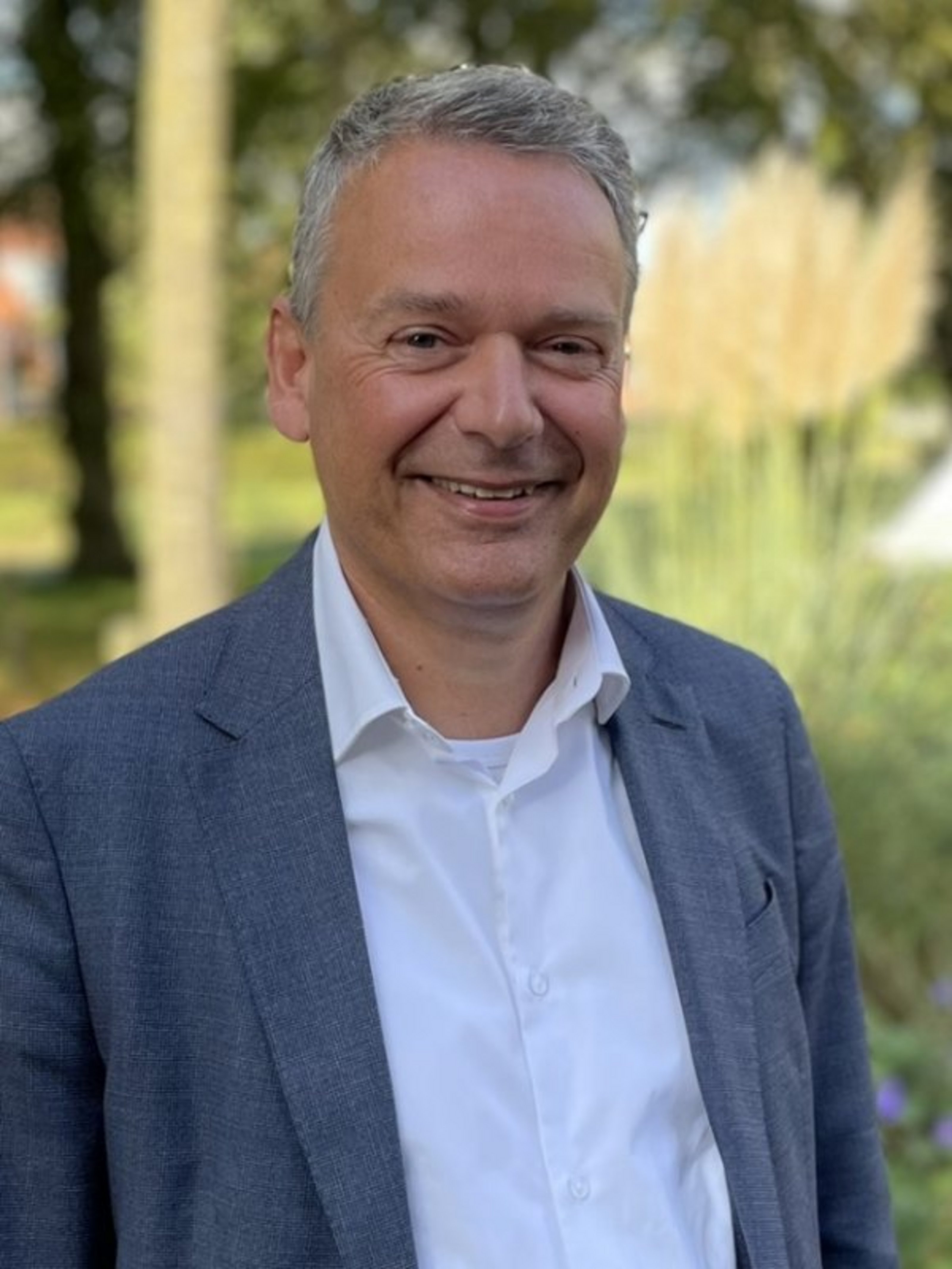 Ein Porträtbild mit dem Ansprechpartner Stephan Beck: Er trägt eine weiße Hemd mit grauem Jacket und steht vor einigen Bäumen mit Pampasgras.