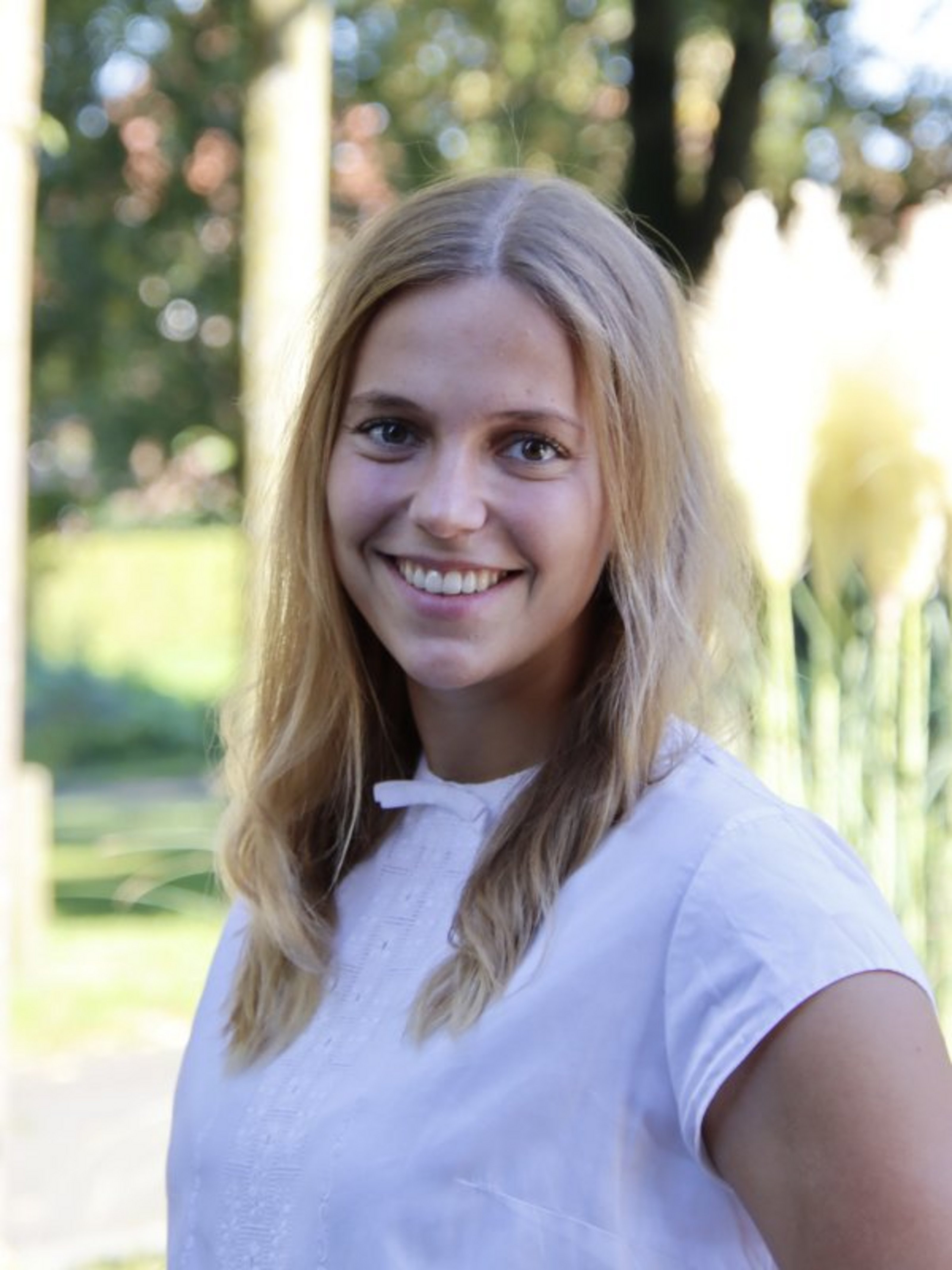 Ein Porträtbild mit der Ansprechpartnerin Carolin Lehmann: Sie trägt eine weiße Bluse und steht vor einigen Bäumen.