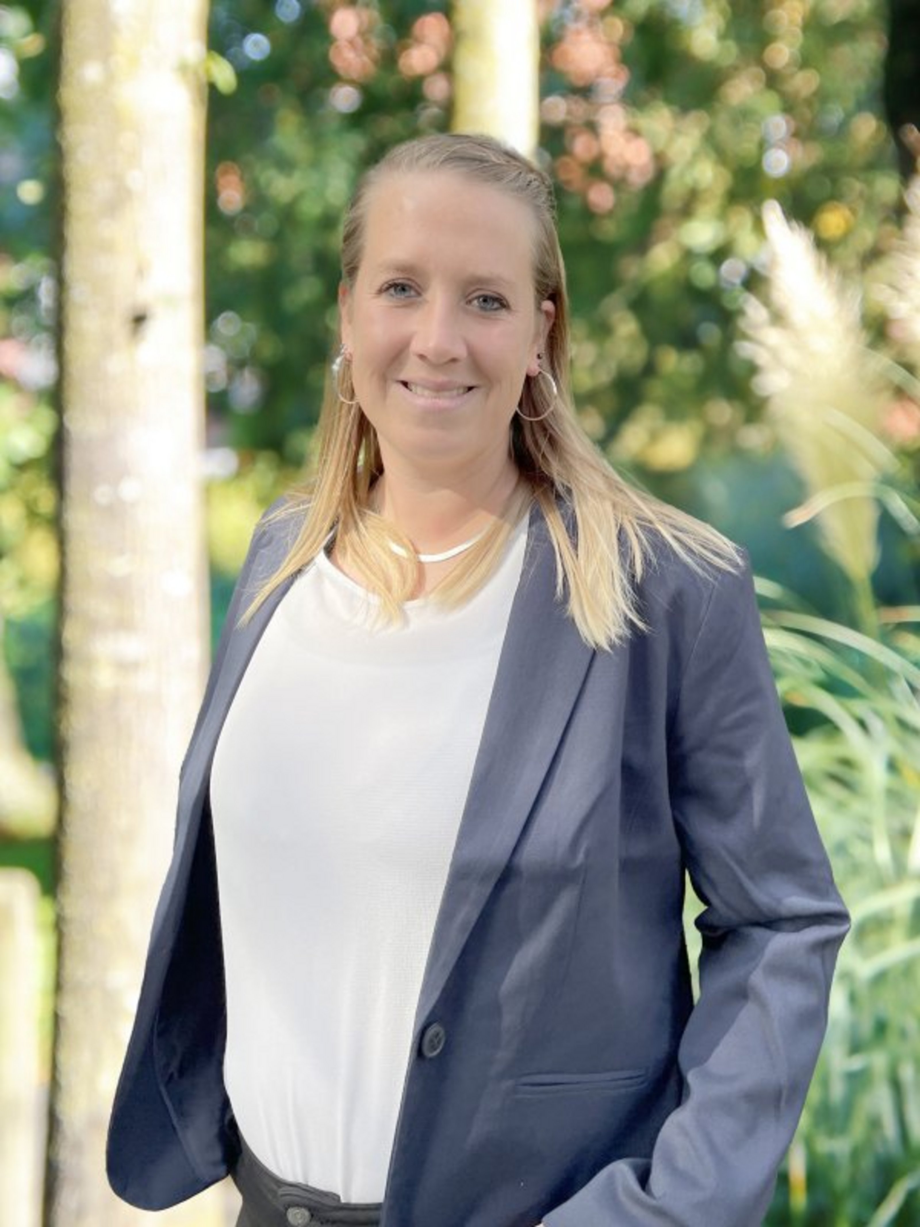 Ein Porträtbild mit der Ansprechpartnerin Johanna Stauvermann: Sie trägt eine weiße Bluse mit grauem Blazer und steht vor einigen Bäumen mit Pampasgras.